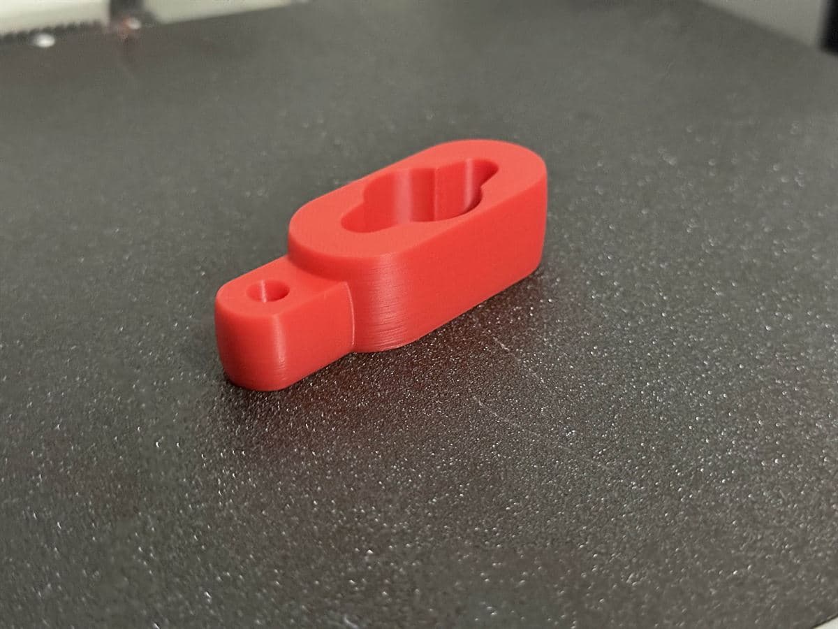 A red PLA+ clamp with a keyhole-shaped slot and a small through-hole, sitting on a textured print bed